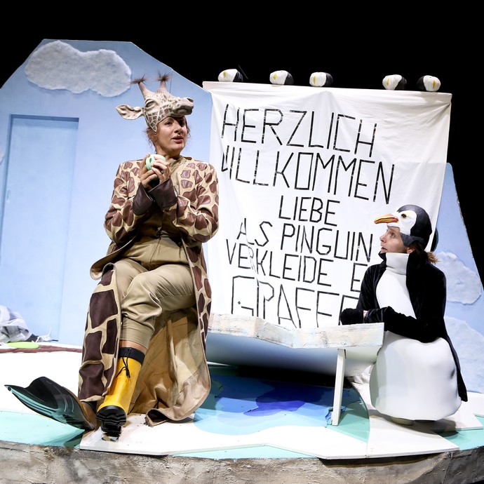 Szene aus "Viele Grüße, deine Giraffe" mit zwei Personen verkleidet als Pinguin und Giraffe. Szene aus "Viele Grüße, deine Giraffe" mit zwei Personen verkleidet als Pinguin und Giraffe.